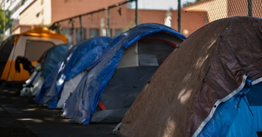 Homeless. poverty, A row of dome tents covered with blue and brown tarps lined up along a shaded sidewalk beside a chain-link fence.