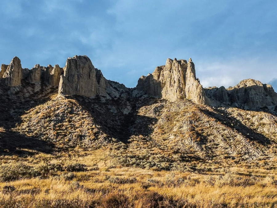 Rocky cliffs and spires rising above a sunlit, grassy hillside under a cloudy blue sky. McAbee fossil beds, Cache Creek, BC